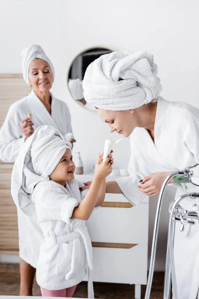 Enfant souriant en peignoir tenant la crème près des parents lesbiennes dans la salle de bain — Photo de stock