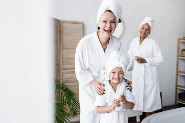 Cheerful woman hugging adopted daughter in bathrobe holding cream in bathroom — Stock Photo