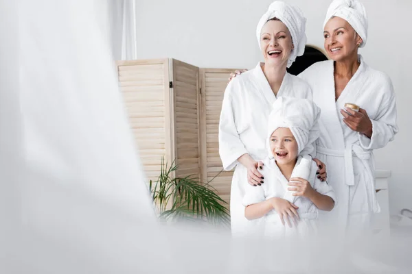 Couple lesbien joyeux et fille adoptive en peignoirs tenant crème cosmétique dans la salle de bain — Photo de stock