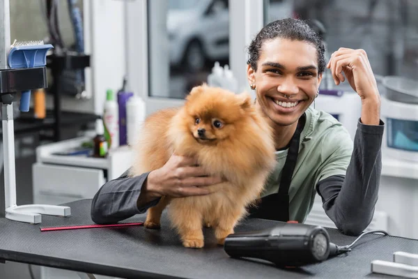 Cheerful african american groomer smiling at camera near spitz and hair dryer on table — Stock Photo