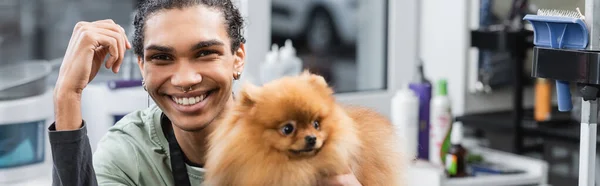 Joyful african american pet hairdresser looking at camera near pomeranian spitz, banner — Stock Photo