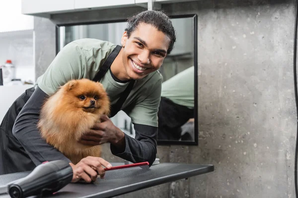 Junge afrikanisch-amerikanische Tierfriseurin lächelt in die Kamera in der Nähe von flauschigem Spitz und Fön auf dem Pflegetisch — Stockfoto