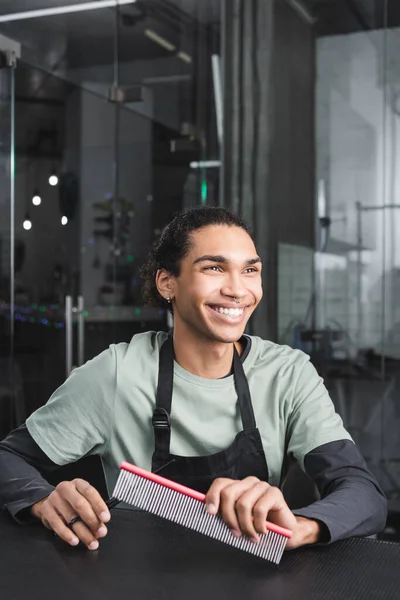 Alegre afroamericano groomer holding peine y tijeras mientras sentado en barbería - foto de stock