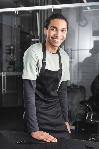 Happy african american hairdresser in apron looking at camera in grooming salon — Stock Photo