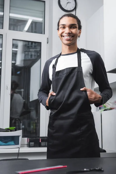 Jovem barbeiro afro-americano em avental sorrindo para a câmera no salão de aliciamento — Fotografia de Stock