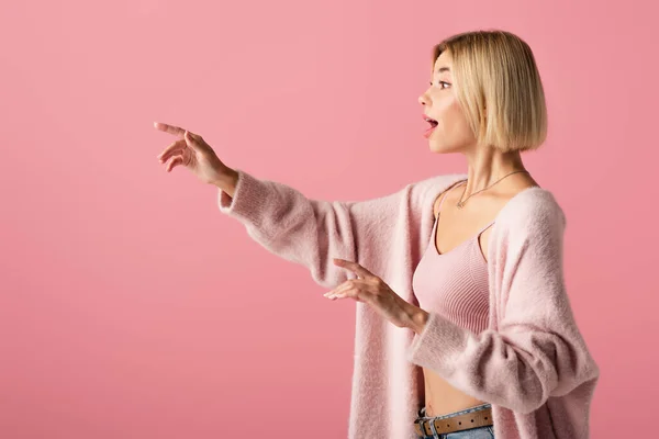 Vista lateral de la joven conmocionada en suave cárdigan mirando hacia otro lado y haciendo un gesto aislado en rosa - foto de stock