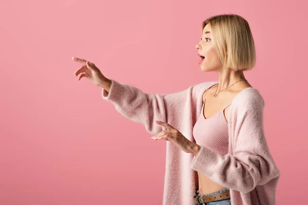 Vue latérale de jeune femme choquée dans un cardigan doux regardant loin et gesticulant isolé sur rose — Photo de stock