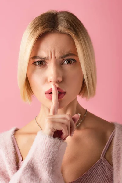 Rubia joven frunciendo el ceño y mostrando el signo de silencio aislado en rosa - foto de stock