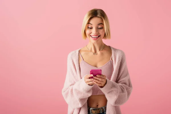 Щаслива молода жінка в м'якому кардигані за допомогою смартфона ізольована на рожевому — стокове фото