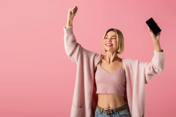 Feliz joven en suave cárdigan sosteniendo teléfono inteligente con pantalla en blanco aislado en rosa - foto de stock