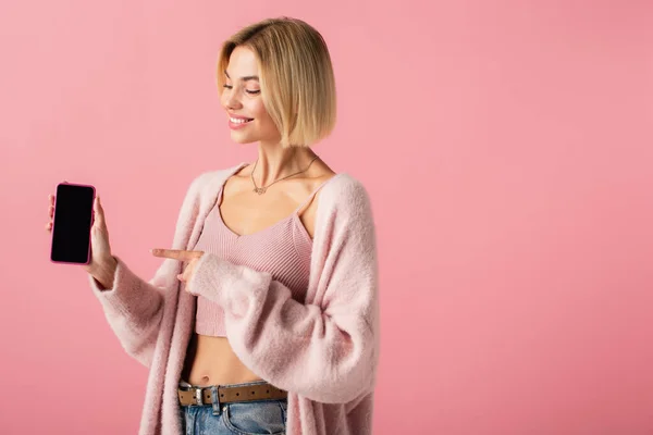 Joyeuse jeune femme en doux cardigan pointant vers le smartphone avec écran vide isolé sur rose — Photo de stock