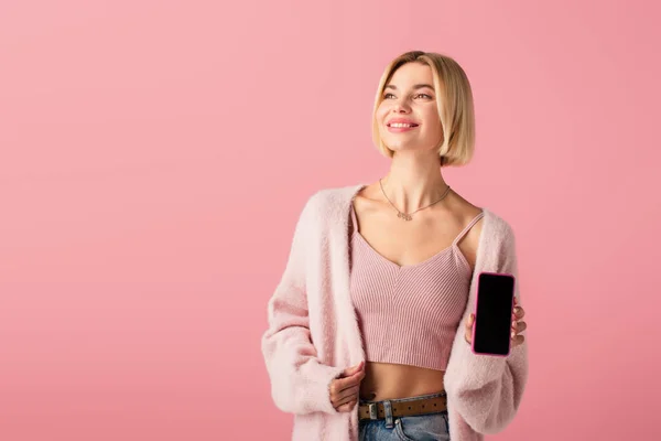 Mujer joven positiva en un suave cárdigan que sostiene el teléfono inteligente con pantalla en blanco aislado en rosa - foto de stock