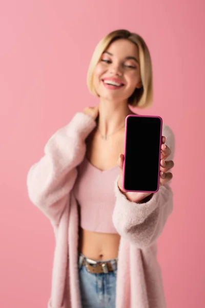 Mujer joven borrosa y feliz en un suave cárdigan que sostiene el teléfono inteligente con pantalla en blanco aislado en rosa - foto de stock