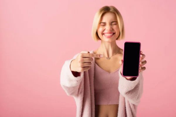 Verschwommen und glücklich junge Frau in weicher Strickjacke zeigt auf Smartphone mit leerem Bildschirm isoliert auf rosa — Stockfoto