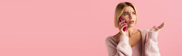 Displeased young woman in soft cardigan talking on smartphone isolated on pink, banner — Stock Photo
