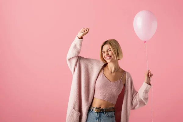 Радостная молодая женщина в кардигане держит воздушный шар, изолированный на розовый — стоковое фото