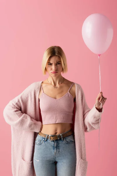 Незадоволена молода жінка в кардигані тримає повітряну кулю і стоїть з рукою на стегні ізольовано на рожевому — стокове фото