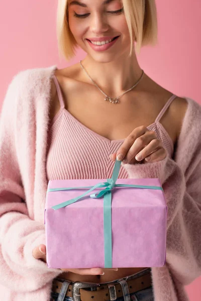 Alegre joven mujer en cárdigan tirando de cinta azul en caja de regalo envuelto aislado en rosa - foto de stock