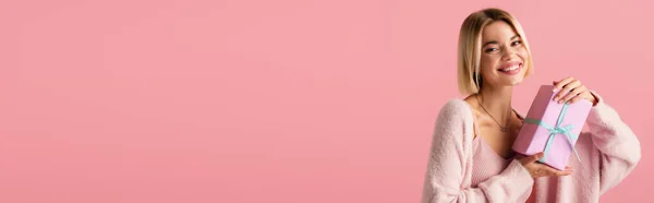 Fröhliche junge Frau in Strickjacke mit eingewickelter Geschenkbox isoliert auf rosa, Banner — Stockfoto