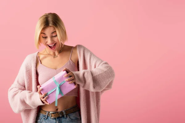 Amazed young woman in cardigan holding wrapped gift box isolated on pink — Stock Photo