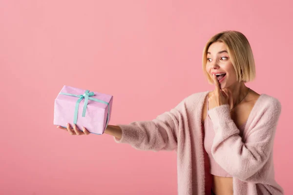 Збуджена молода жінка в кардигані, що закриває рот пальцем, тримаючи загорнуту подарункову коробку ізольовану на рожевому — стокове фото
