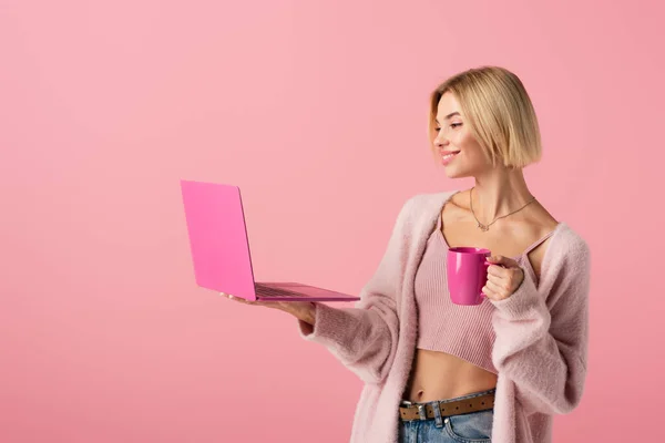 Щаслива жінка тримає кухоль з кавою і ноутбуком ізольовано на рожевому — стокове фото