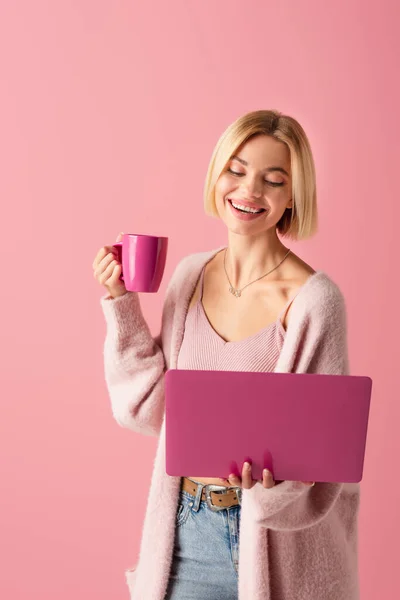 Mujer alegre sosteniendo taza con café y portátil aislado en rosa - foto de stock