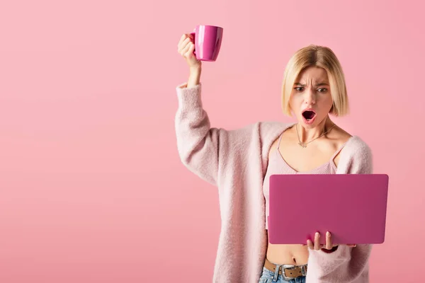 В шоке женщина держит кружку с кофе и ноутбук изолирован на розовый — стоковое фото