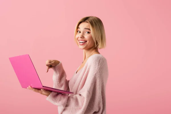 Happy young woman pointing with finger at laptop isolated on pink — Stock Photo