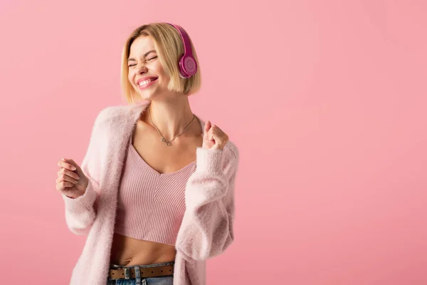 Allegra donna bionda che ascolta musica in cuffie wireless isolate su rosa — Foto stock