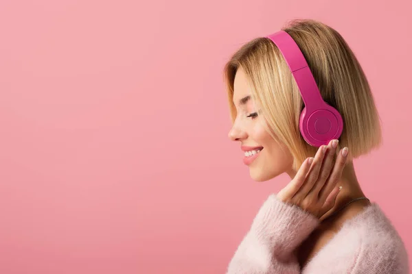 Вид сбоку жизнерадостной блондинки, слушающей музыку в беспроводных наушниках, изолированных на розовом фоне — стоковое фото