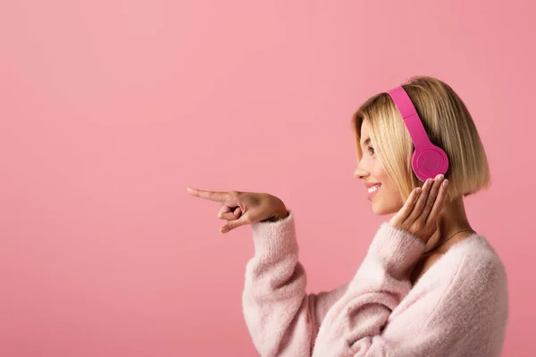 Seitenansicht einer fröhlichen blonden Frau, die Musik in drahtlosen Kopfhörern hört und mit dem Finger isoliert auf rosa zeigt — Stockfoto