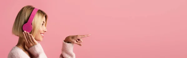 Vue latérale de femme joyeuse écoutant de la musique dans des écouteurs sans fil et pointant avec le doigt isolé sur rose, bannière — Photo de stock