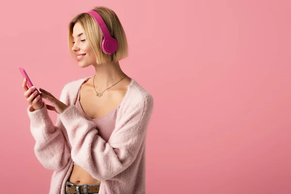 Mujer alegre escuchando música en auriculares inalámbricos y utilizando un teléfono inteligente aislado en rosa - foto de stock