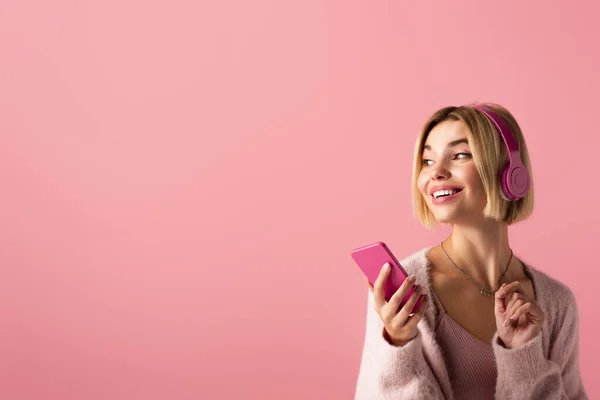 Mujer alegre escuchando música en auriculares inalámbricos y sosteniendo teléfono inteligente aislado en rosa - foto de stock