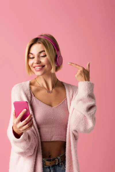 Femme gaie pointant vers les écouteurs sans fil et tenant smartphone isolé sur rose — Photo de stock