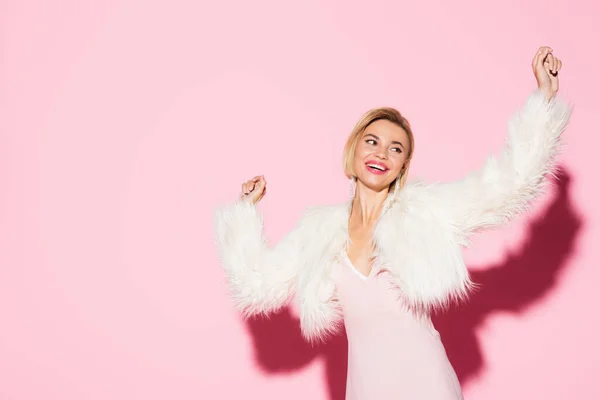 Excited blonde woman in slip dress and faux fur jacket on pink — Stock Photo