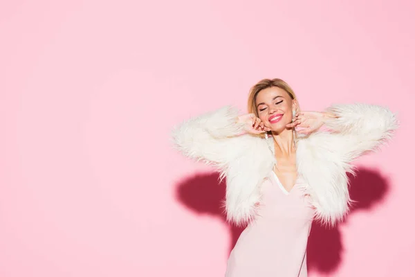 Joyful blonde woman in slip dress and faux fur jacket on pink — Stock Photo
