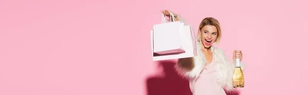 Femme étonnée en robe glissante et veste en fausse fourrure tenant des sacs à provisions et une bouteille de champagne sur rose, bannière — Photo de stock