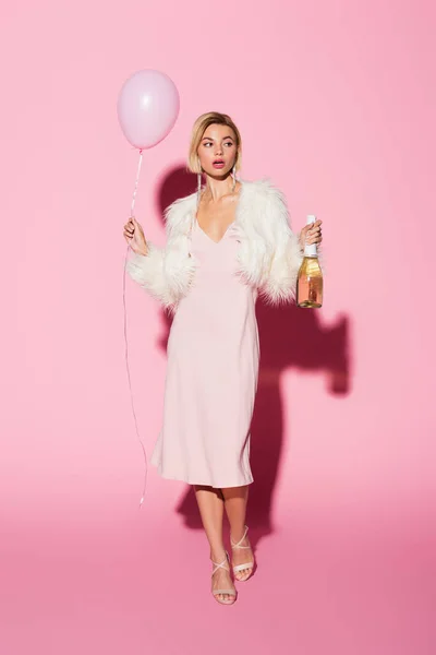 Full length of shocked woman in slip dress and faux fur jacket holding balloon and bottle of champagne on pink — Stock Photo