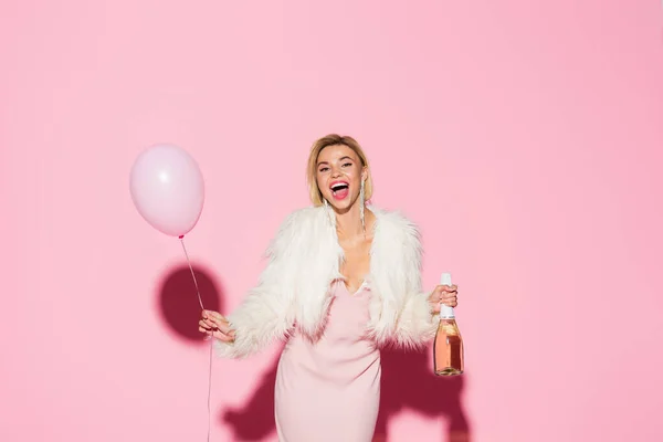 Donna eccitata in abito antiscivolo e giacca di pelliccia sintetica con palloncino e bottiglia di champagne su rosa — Foto stock