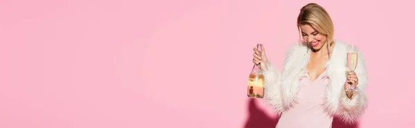 Femme blonde souriante en robe slim et veste en fausse fourrure tenant bouteille de champagne et verre sur rose, bannière — Photo de stock