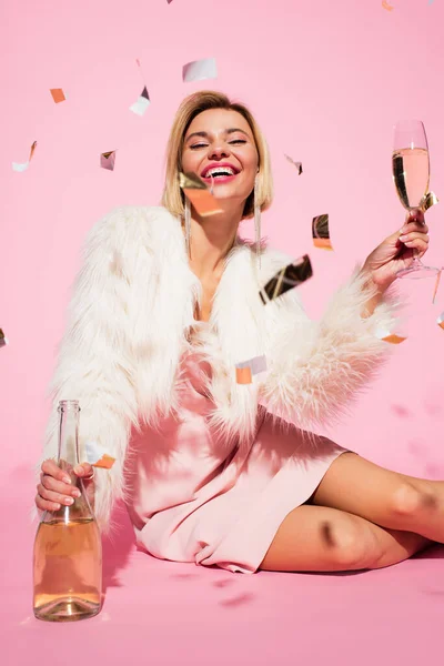 Positive Frau in Slip-Kleid und Kunstpelzjacke hält Flasche Champagner und Glas in der Nähe fallenden Konfetti auf rosa — Stockfoto