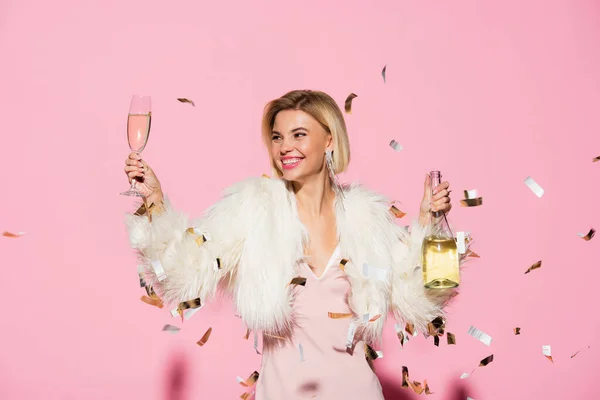 Cheerful woman in slip dress and faux fur jacket holding bottle of champagne and glass near falling confetti on pink — Stock Photo