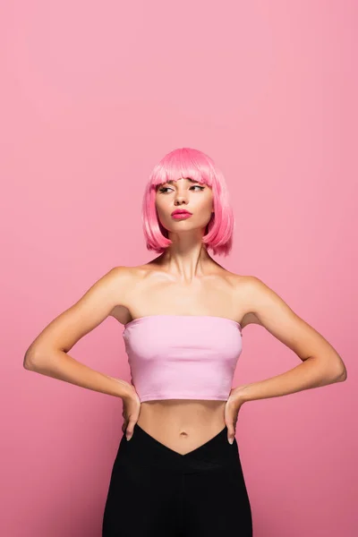 Giovane modella con frangia e capelli colorati in posa con le mani sui fianchi isolate su rosa — Foto stock