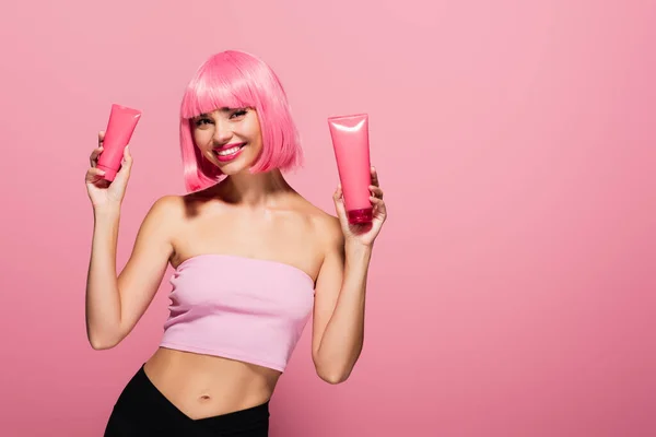 Jovem feliz com cabelos coloridos e franja segurando tubos com loção isolada em rosa — Fotografia de Stock