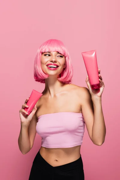 Alegre joven con el pelo coloreado y flequillo sosteniendo tubos con loción aislada en rosa - foto de stock