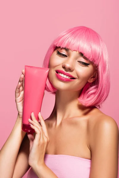 Alegre joven mujer con el pelo de color y flequillo sosteniendo tubo con loción aislada en rosa - foto de stock