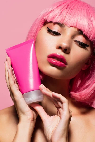 Close up de jovem mulher com cabelos coloridos e franja segurando tubo com loção isolada em rosa — Fotografia de Stock