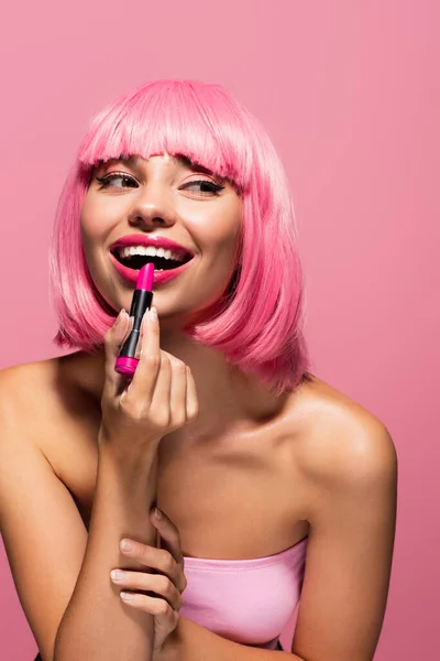 Cheerful young woman with colored hair and bare shoulders applying lipstick isolated on pink — Stock Photo